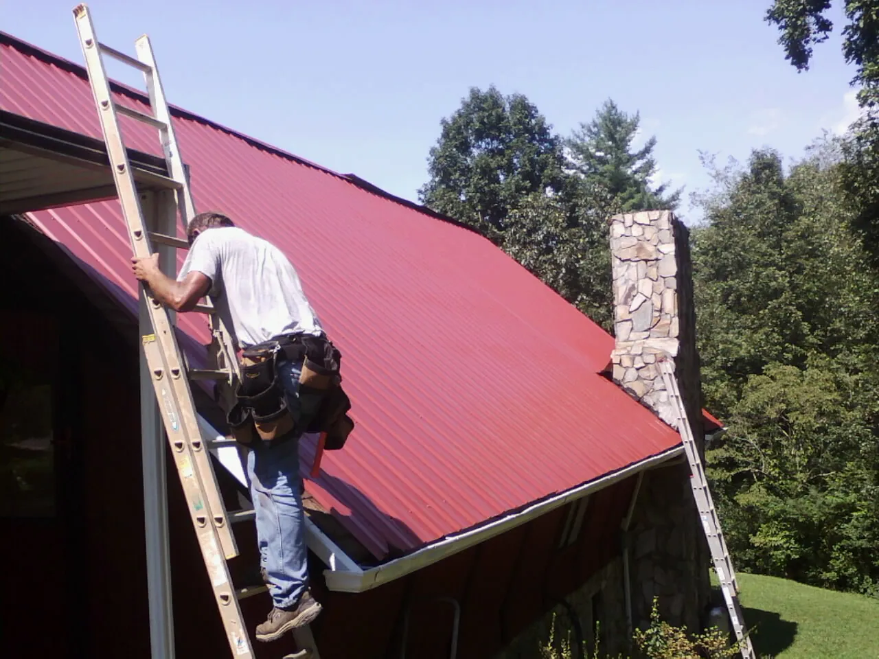 Roofer on a ladder beside a red metal roof and stone chimney — Soffit & Fascia Repair by Roofing Brothers in Plymouth Meeting